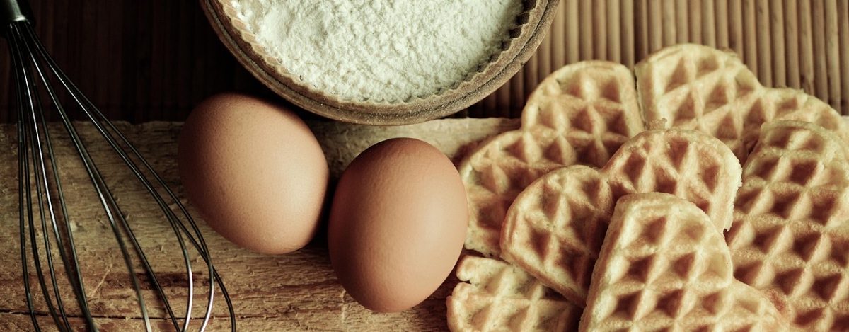 Ägg, vetemjöl i en skål, en visp och flera nygräddade våfflor på ett träunderlag.